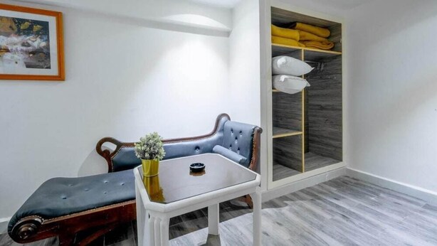 Una habitación con un banco azul acolchado, una mesita con un florero, un armario con estantes y un cuadro en la pared.
