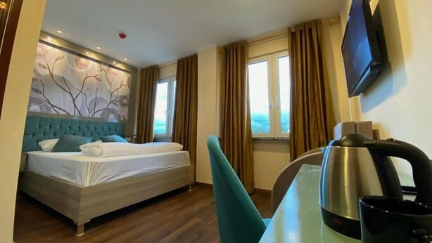 A hotel room with a bed, a desk with a kettle, a chair, a TV, and a window with curtains. - Hotel Grand Inn (Berlin)