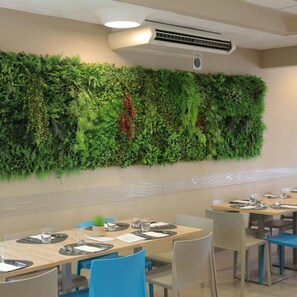 A restaurant with a green vertical garden wall, tables with blue chairs, and a ceiling air conditioning unit. - Hotel Sole (Cattolica)