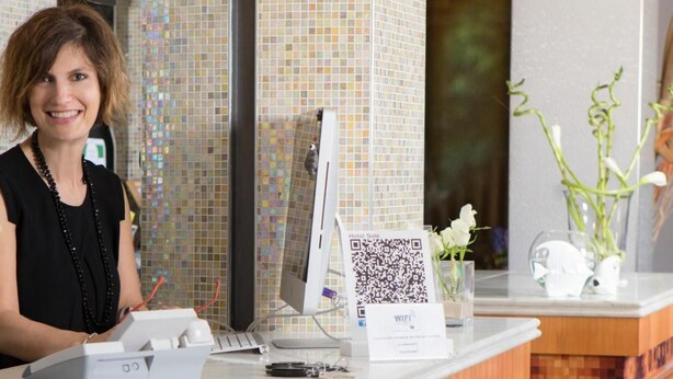 A hotel reception desk with a computer, a QR code for WiFi, and a vase with white flowers. - Hotel Sole (Cattolica)
