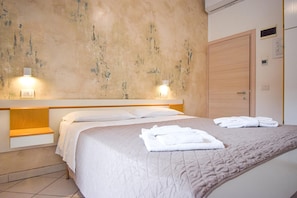 Ein ordentlich bezogenes Bett mit weißen Laken und einer beigen Steudecke, ein hölzerner Kopfteil sowie zwei Wandleuchten.