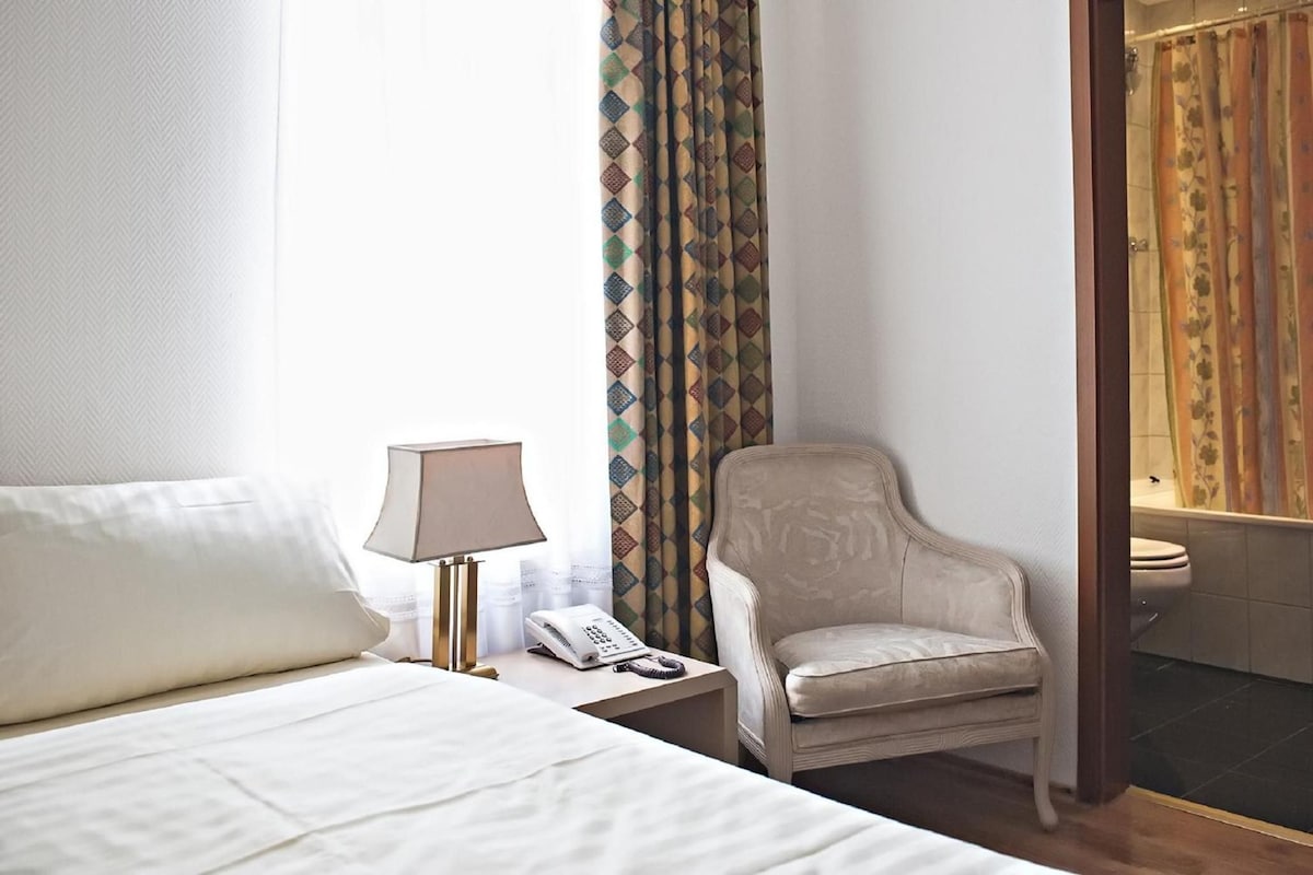 A hotel room with a bed, a chair, a lamp, a telephone, and a bathroom visible through an open door.