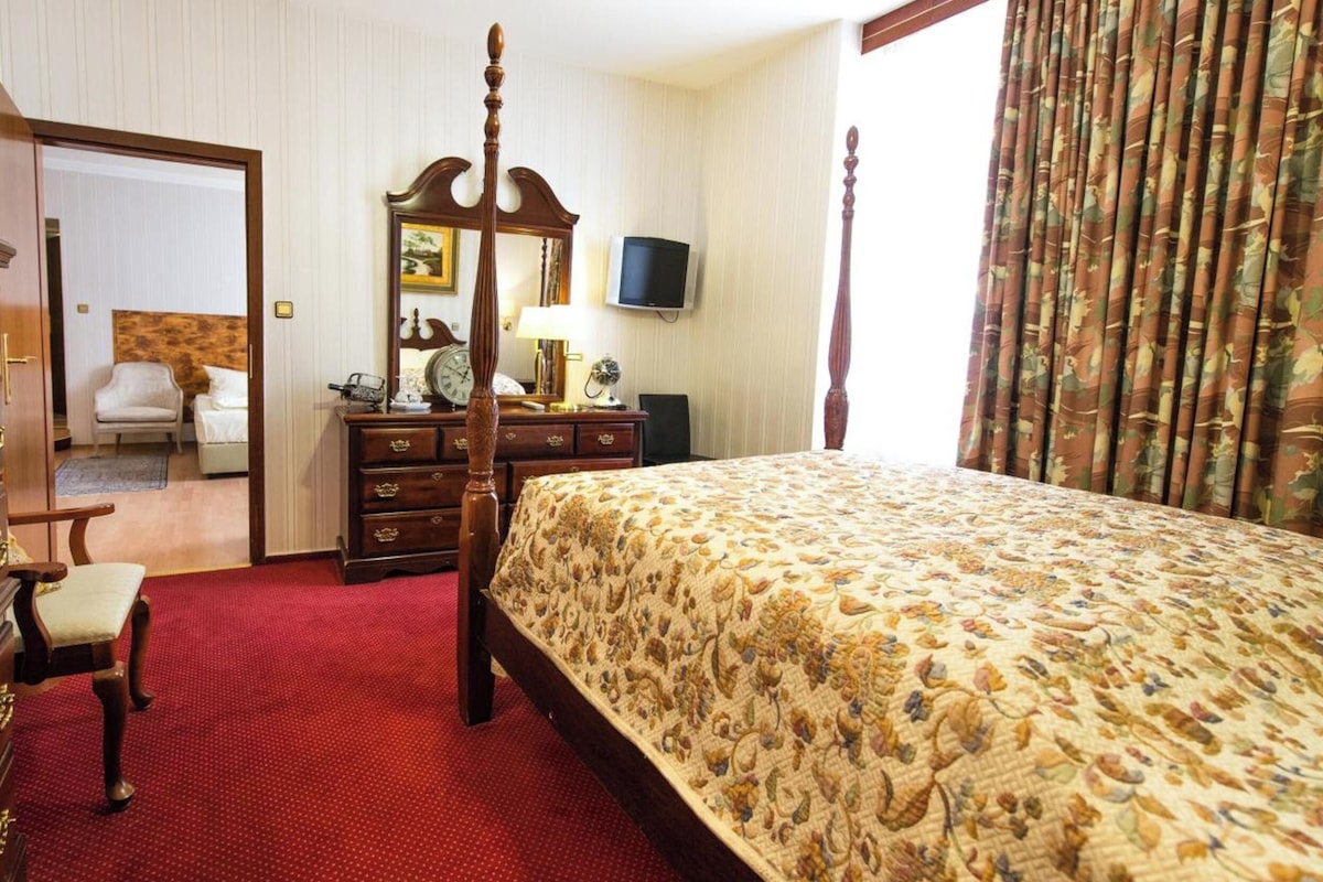 A hotel room with a four-poster bed, a wooden dresser, a television, and a large window with curtains.