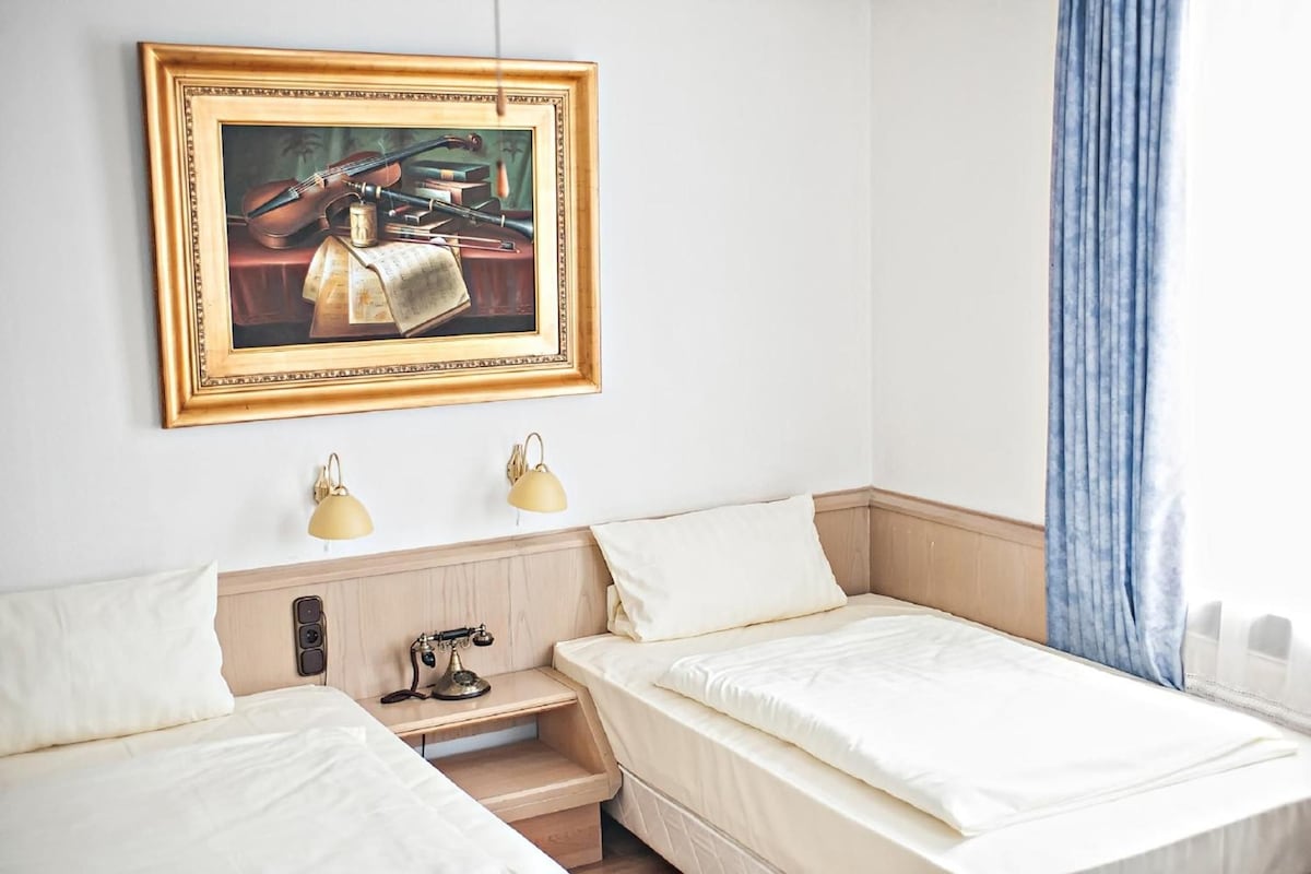 A hotel room with two single beds, a nightstand with a lamp, a painting on the wall, and a window with curtains.