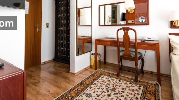 Habitación de hotel con escritorio de madera, silla y un espejo grande. Hay un televisor montado en la pared y una alfombra estampada en el suelo.