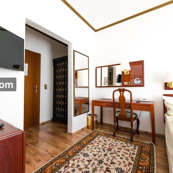 Habitación de hotel con escritorio de madera, silla y un espejo grande. Hay un televisor montado en la pared y una alfombra estampada en el suelo.