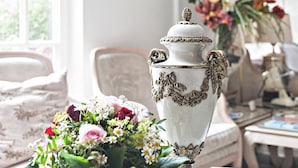 Una habitación con un ventanal grande, un florero blanco con diseños florales y un espejo que refleja la habitación.