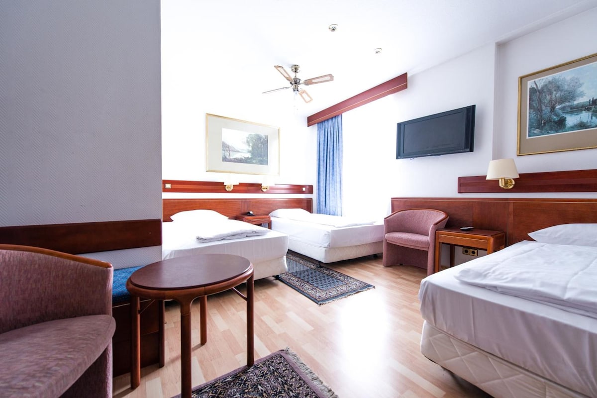 Habitación de hotel con dos camas, un escritorio, una silla, una mesita, ventilador de techo, televisor y ventana con cortinas.