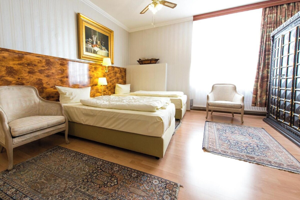 Habitación de hotel con dos camas, un sofá, una silla, un ventilador de techo, un cuadro y un ventanal con cortinas.