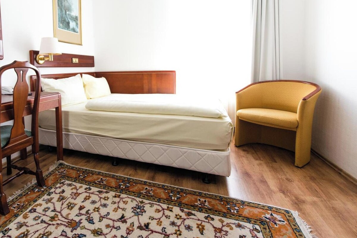 Una habitación de hotel con una cama, una silla, un escritorio, una lámpara y una alfombra.