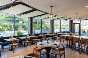 Um restaurante com mesas redondas e cabines, amplas janelas com vista para o exterior e iluminação pendente.