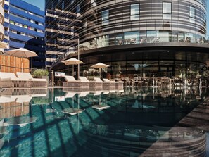 Um hotel moderno com piscina, espreguiçadeiras, guarda-sóis e o reflexo da arquitetura do edifício.