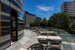 Uma área externa com mesas e cadeiras, guarda-corpo de vidro e um edifício de design moderno.