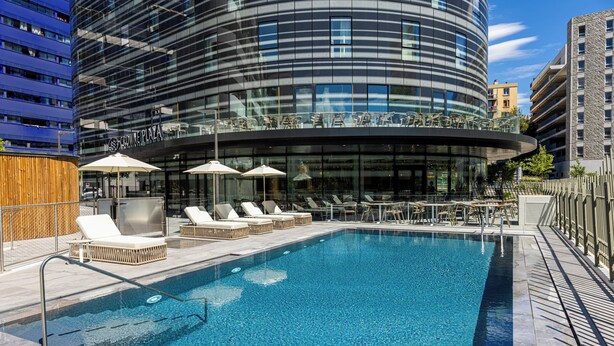 Un hotel moderno con fachada de vidrio curva, un bar en la azotea con mesas al aire libre y una piscina con tumbonas.