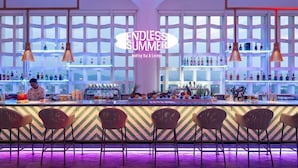 A bar area with a chevron-patterned wall, multiple hanging pendant lights, and a bartender at the counter. - Wink Hotel Danang Riverside (Da Nang)
