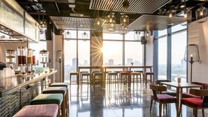 A modern bar area with a high ceiling, exposed ductwork, and large windows offering a city view. - Wink Hotel Danang Riverside (Da Nang)