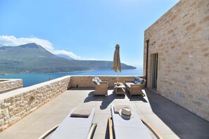Una terraza con muros de piedra, sillones de descanso y una sombrilla, con vistas a un cuerpo de agua y una montaña.