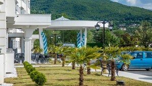 A hotel entrance with a white facade, outdoor seating, palm trees, and a mountain in the background. - Unique Resort & Spa (Ohrid)