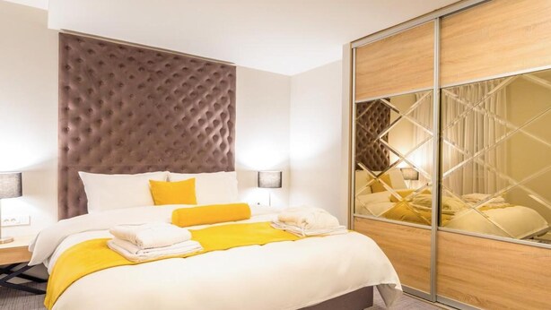 A modern hotel room with a large bed, a tufted headboard, a mirror with a geometric pattern, and a wooden wardrobe. - Unique Resort & Spa (Ohrid)