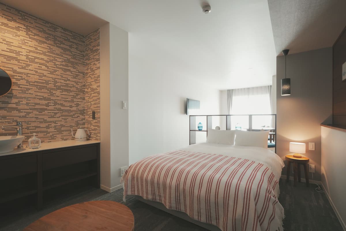 Une chambre d’hôtel avec un mur décoré d’un motif en briques, un lit recouvert d’une courtepointe à rayures, une table de chevet avec une lampe, une fenêtre avec des rideaux et une petite table ronde.