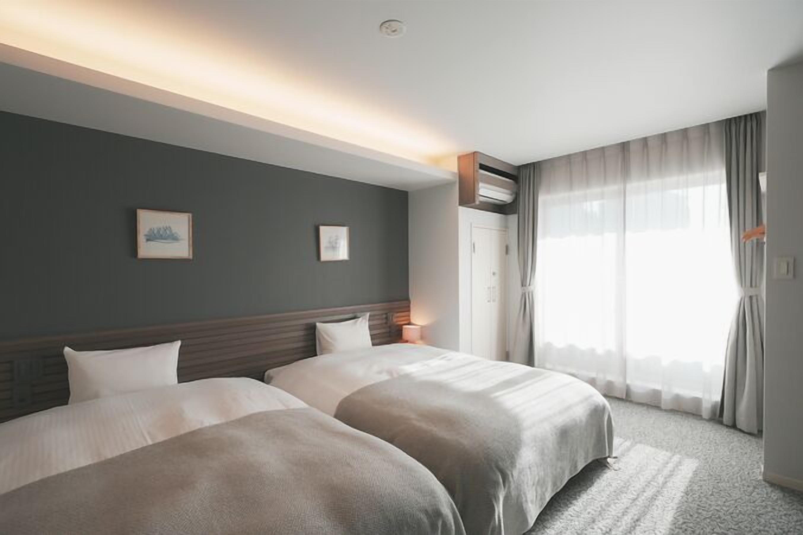 Une chambre d’hôtel avec deux lits, une grande fenêtre avec des rideaux, un luminaire au plafond et un climatiseur fixé au mur.