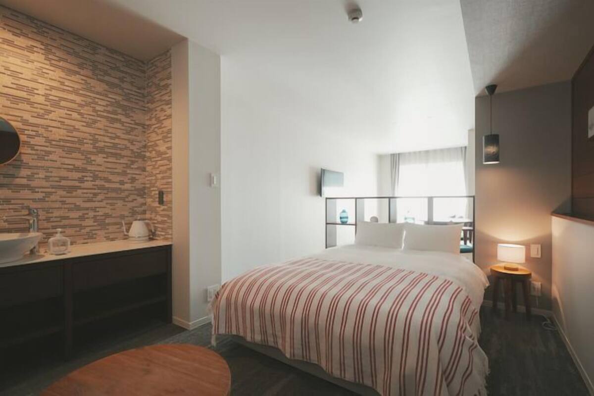 Une chambre d’hôtel avec un mur dont le motif rappelle la pierre, un lit avec une literie à rayures, une table de chevet avec une lampe et une fenêtre avec des rideaux.