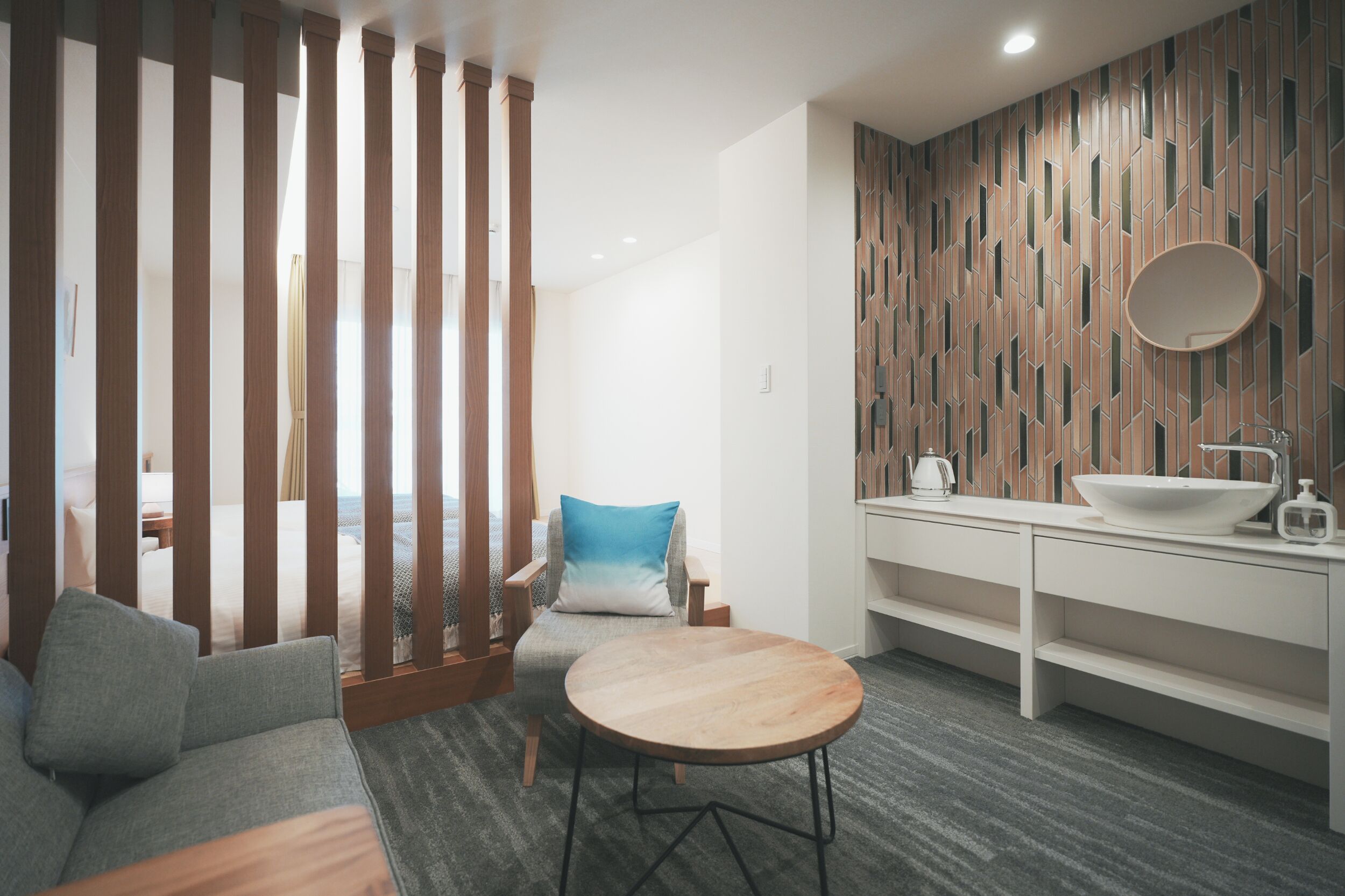 Une chambre d’hôtel moderne avec une cloison en bois, une table ronde, un canapé, un lit, une salle de bain avec un lavabo et un miroir, et un mur décoré d’un motif.