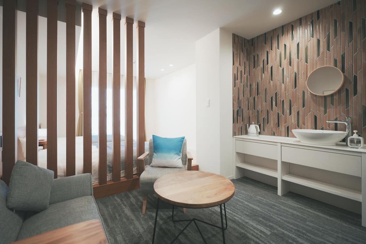 Une chambre d’hôtel moderne avec une cloison en bois, une table ronde, un canapé, un lit, une salle de bain avec un lavabo et un miroir, et un mur décoré d’un motif.