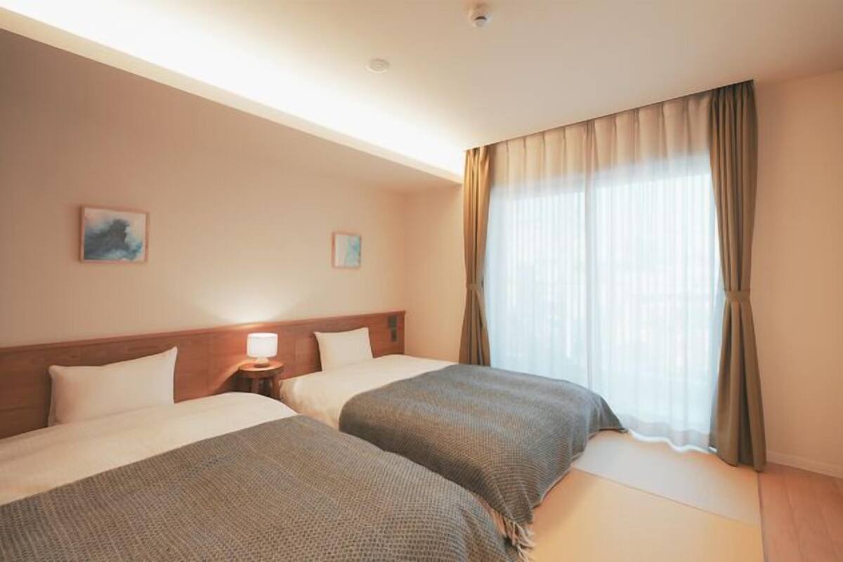 Une chambre d’hôtel avec deux lits, une table de chevet avec une lampe, une grande fenêtre avec des rideaux et des œuvres d’art accrochées aux murs.