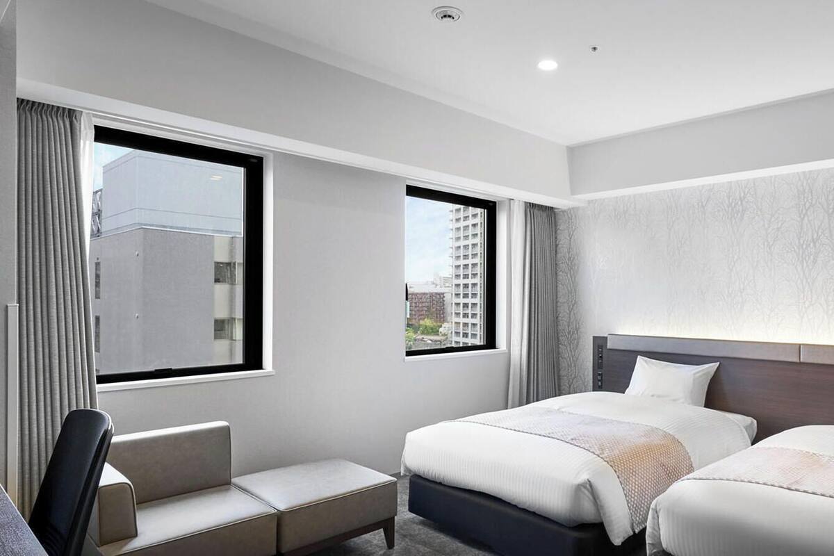 A hotel room with two beds, a seating area, and a large window with a view of buildings.