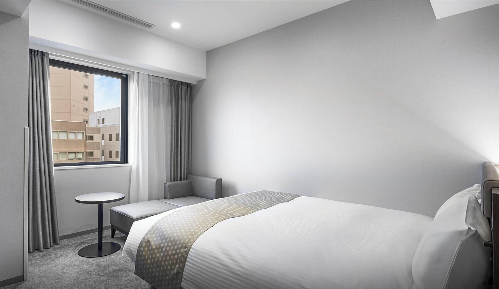 A hotel room with a bed, a small table, a chair, and a window with curtains.