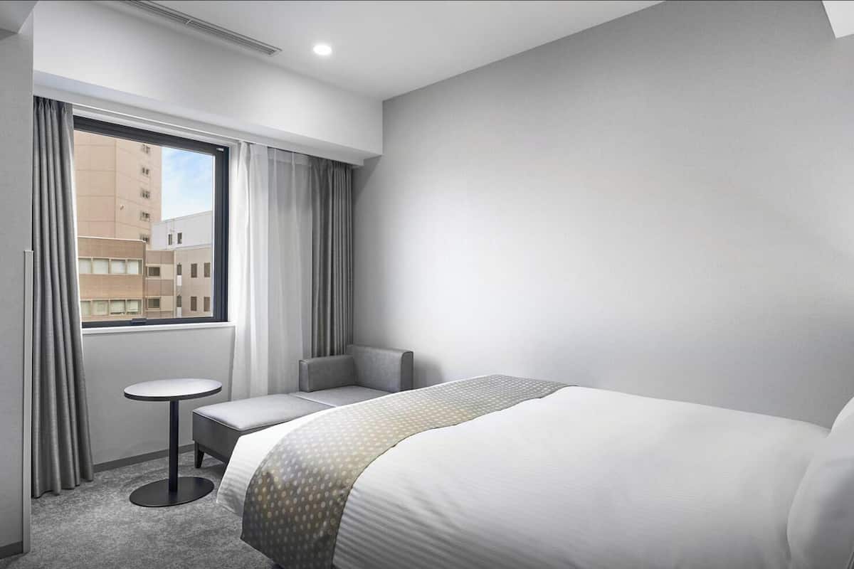 A hotel room with a bed, a small table, a chair, and a window with curtains.