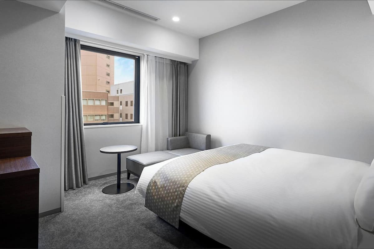 A hotel room with a bed, a small table, a chair, and a window with curtains.