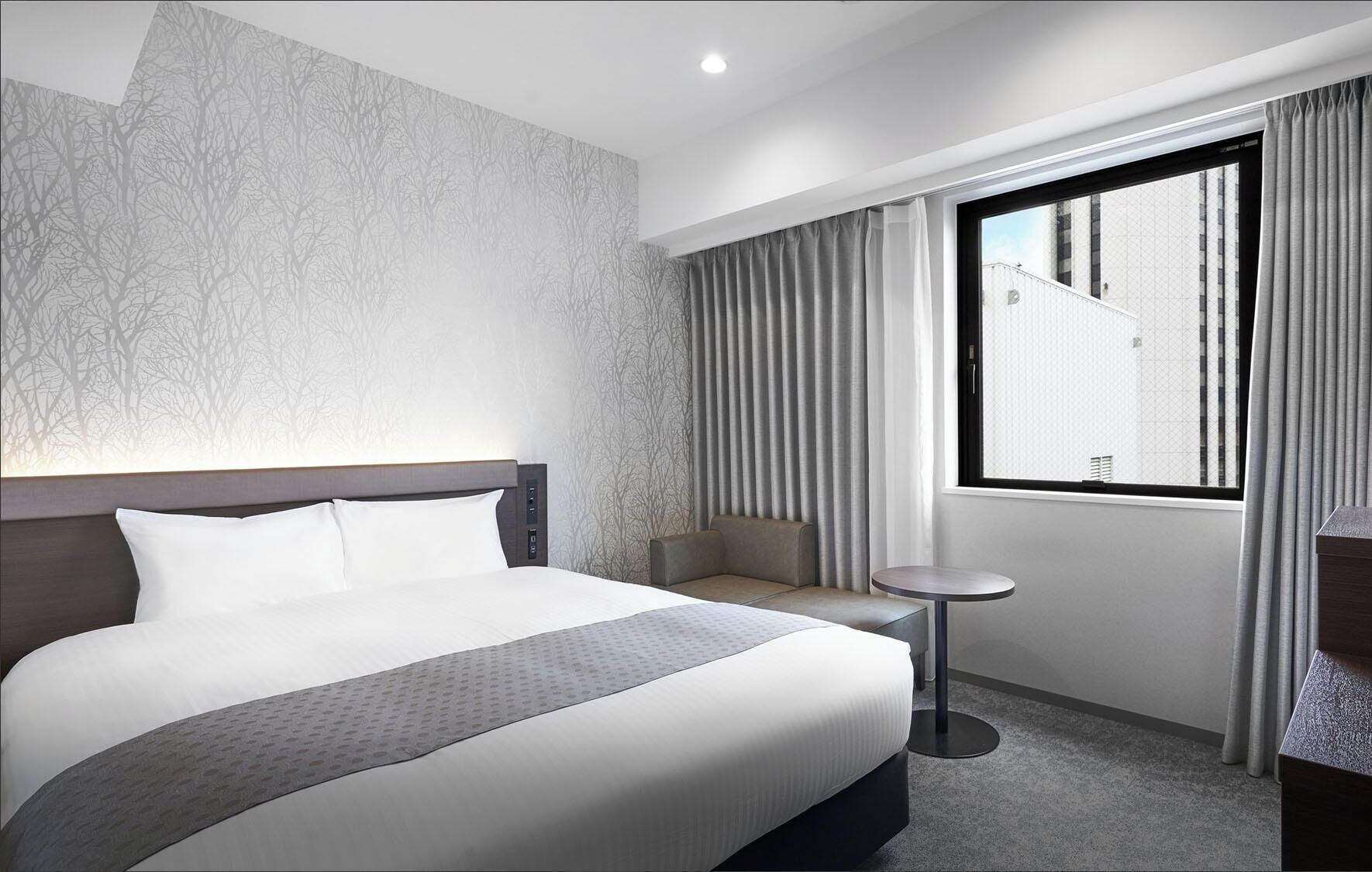 A hotel room with a bed, a small table, a seating area, and a window with curtains.