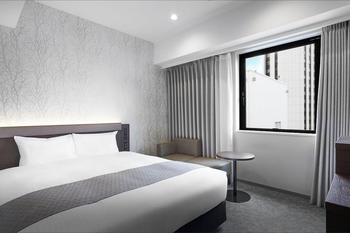 A hotel room with a bed, a small table, a seating area, and a window with curtains.