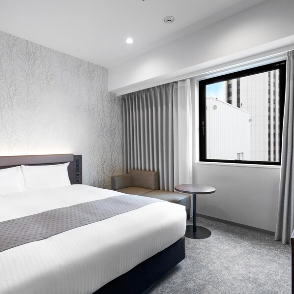 Ein modernes Hotelzimmer mit einem großen Bett, einer kleinen Sitzecke, einem runden Tisch und einem Fenster mit Vorhängen.