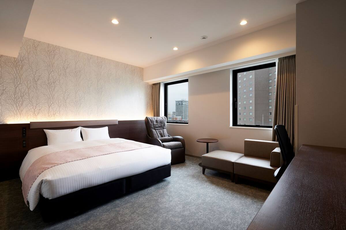 A hotel room with a large bed, a desk, a chair, a small table, and a window with a view of a building.