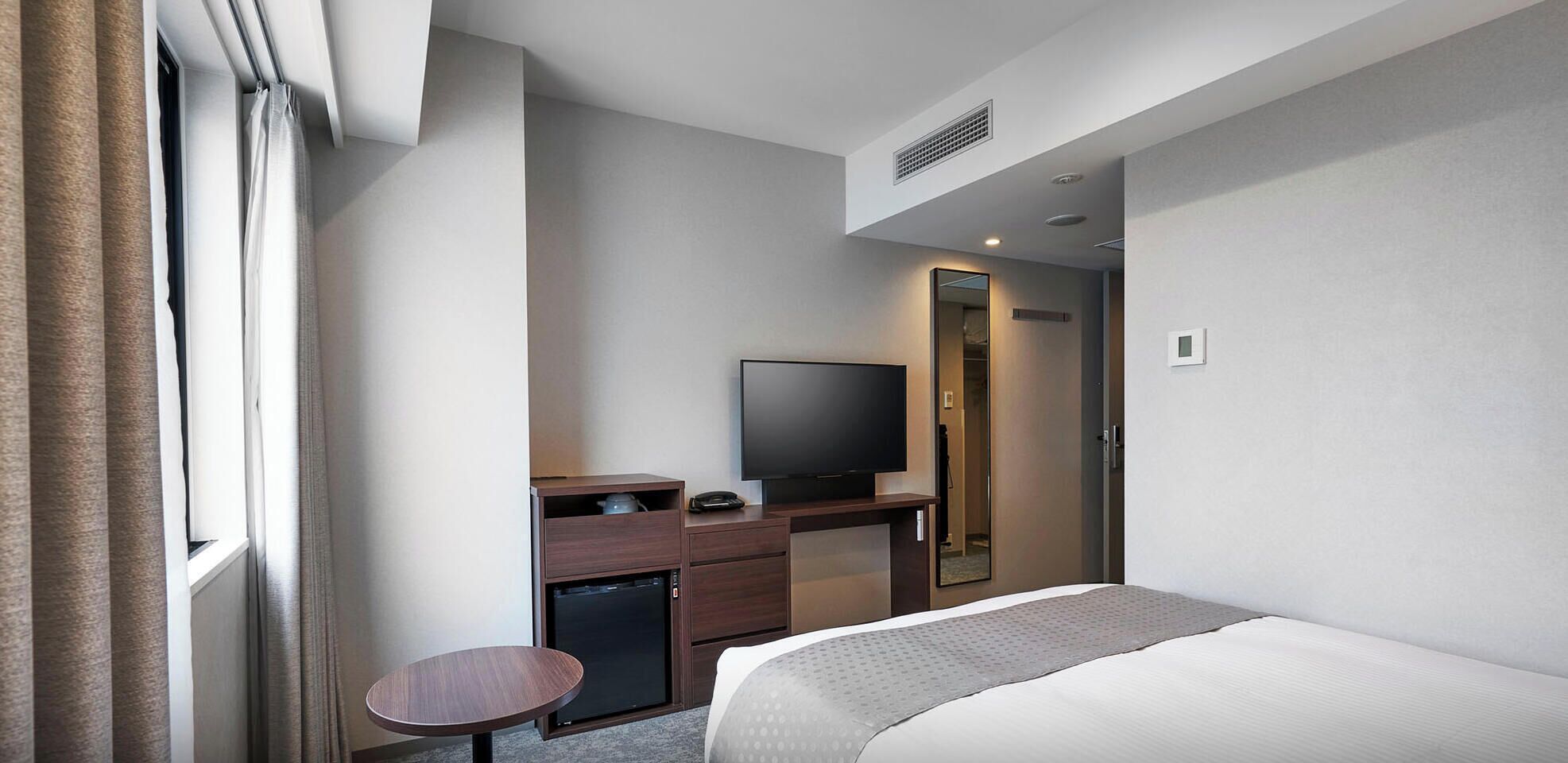 A hotel room with a bed, a desk with a TV, a small table, and a mirror.
