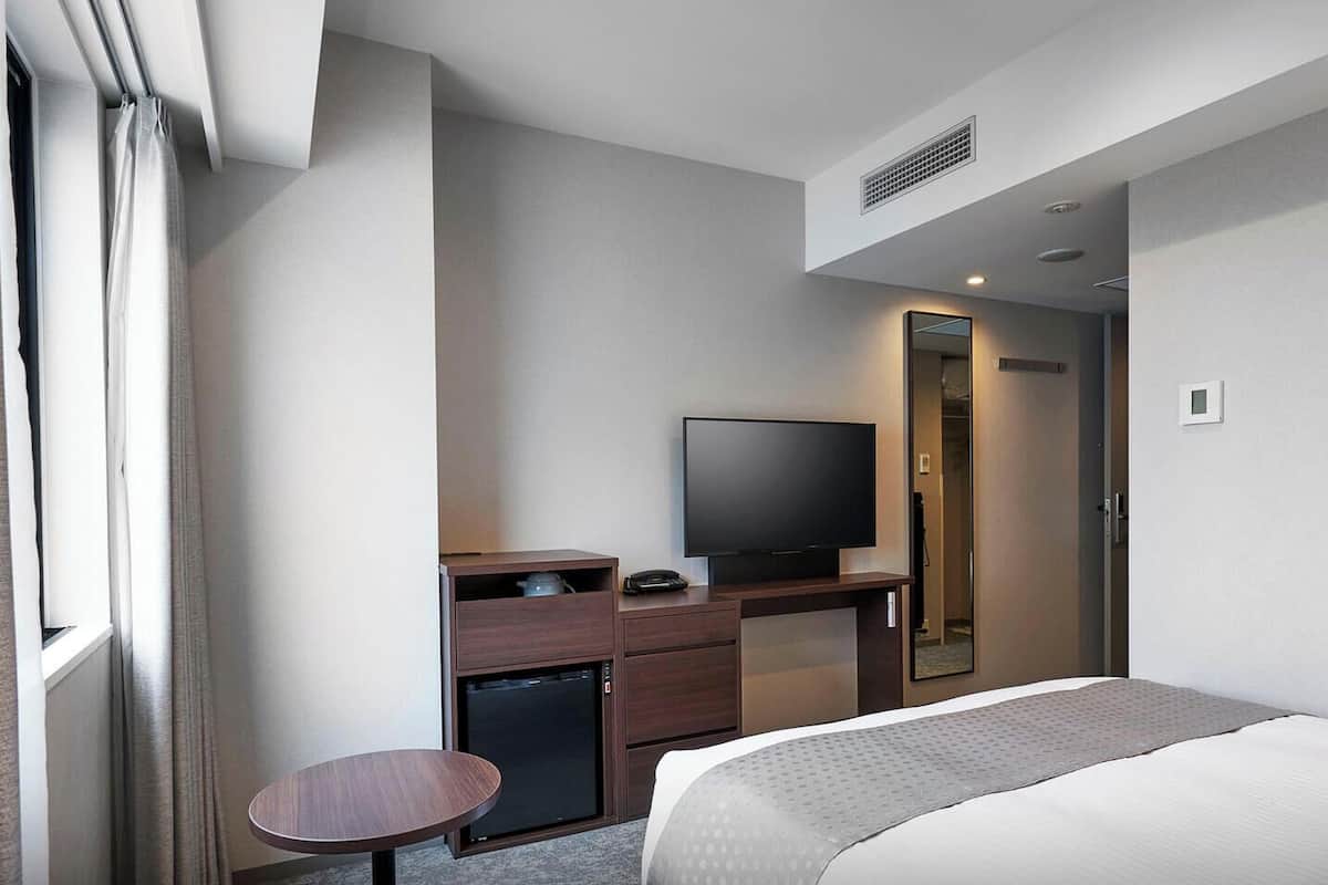 A hotel room with a bed, a desk with a TV, a small table, and a mirror.