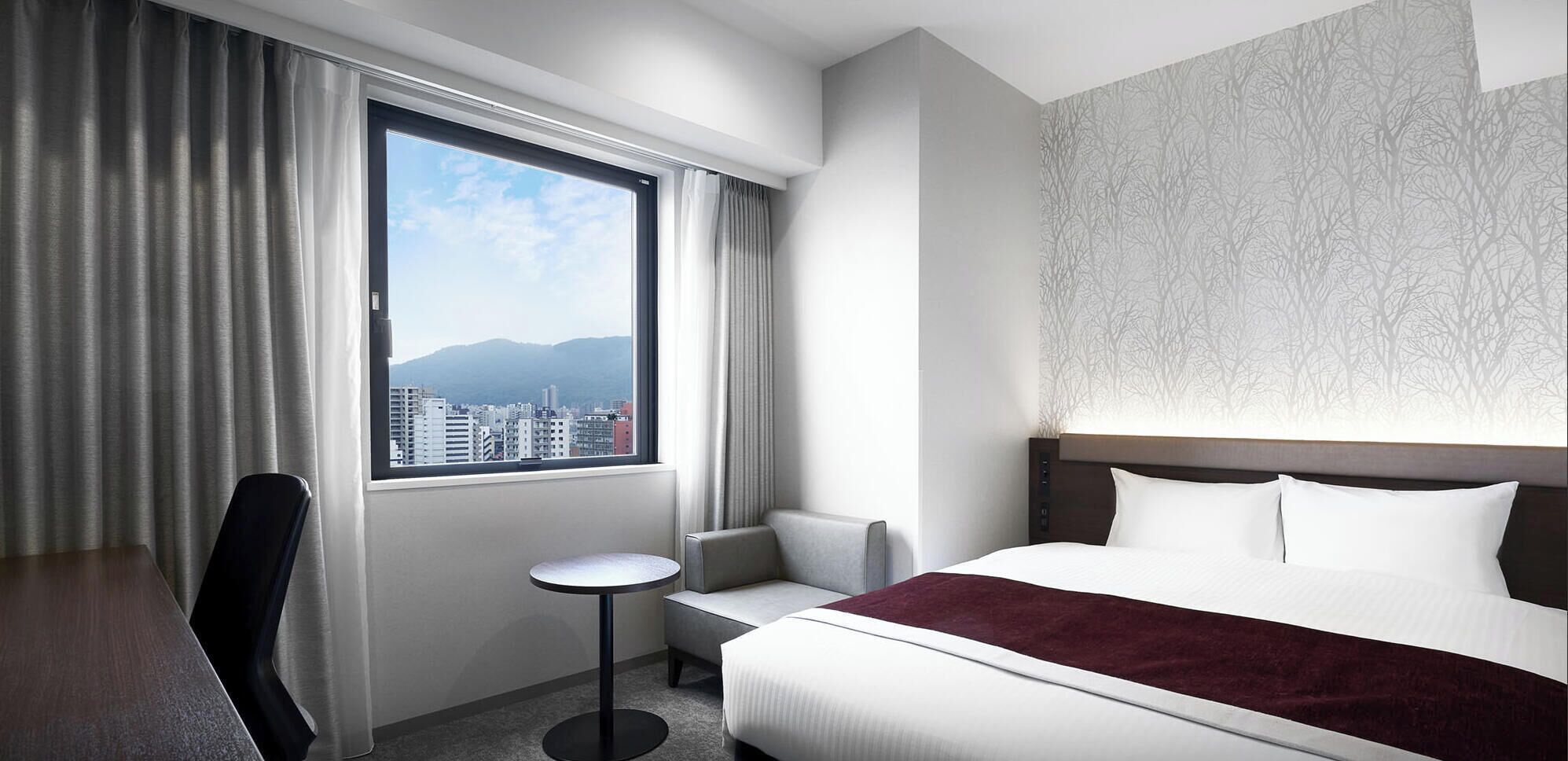 A hotel room with a bed, a chair, a small table, and a view of the cityscape through a large window.