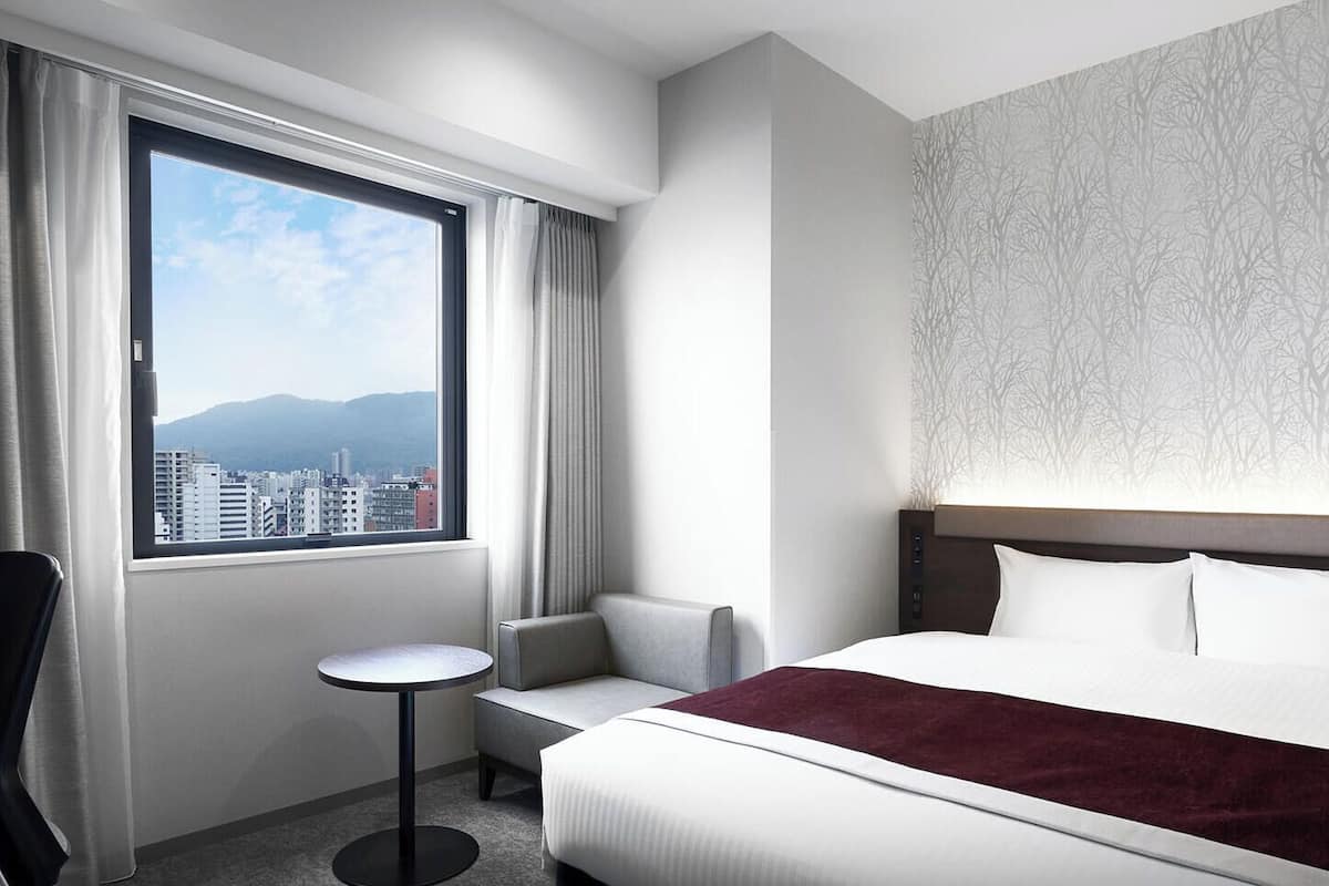 A hotel room with a bed, a chair, a small table, and a view of the cityscape through a large window.