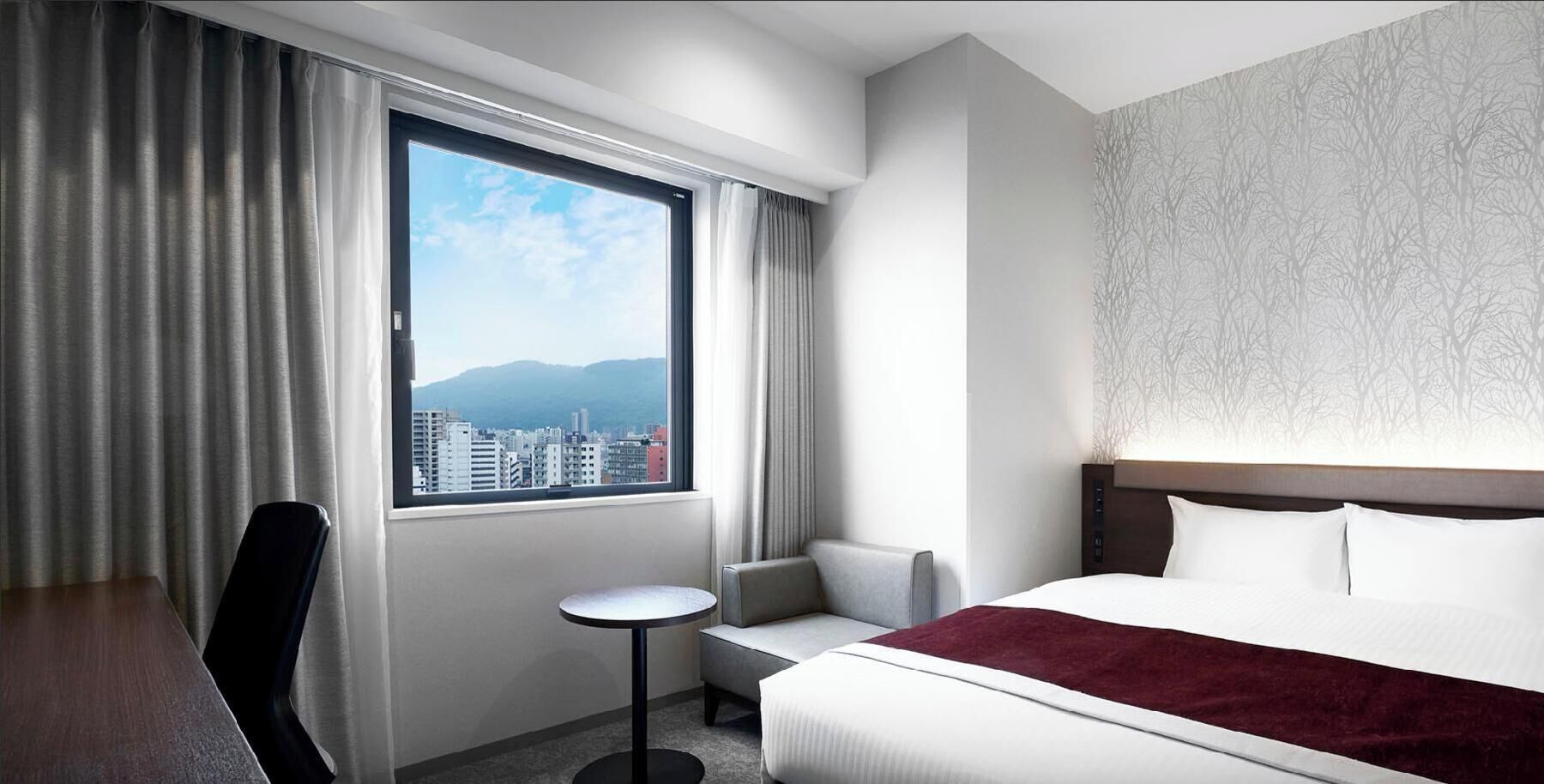 A hotel room with a bed, a desk, a chair, a small table, and a window with a city view.
