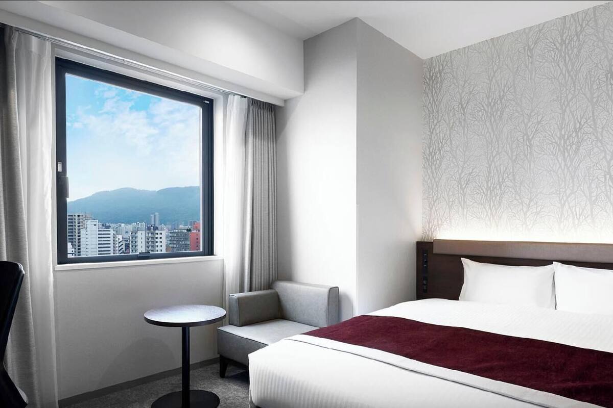 A hotel room with a bed, a desk, a chair, a small table, and a window with a city view.