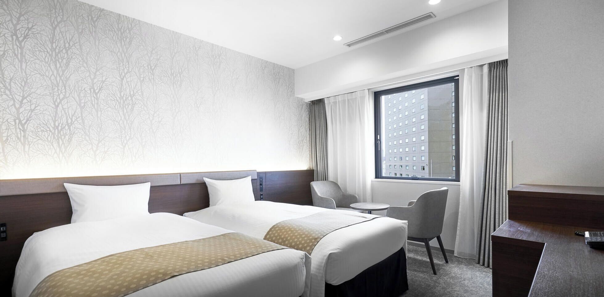 A hotel room with two beds, a desk, a chair, a window with curtains, and a patterned wall.