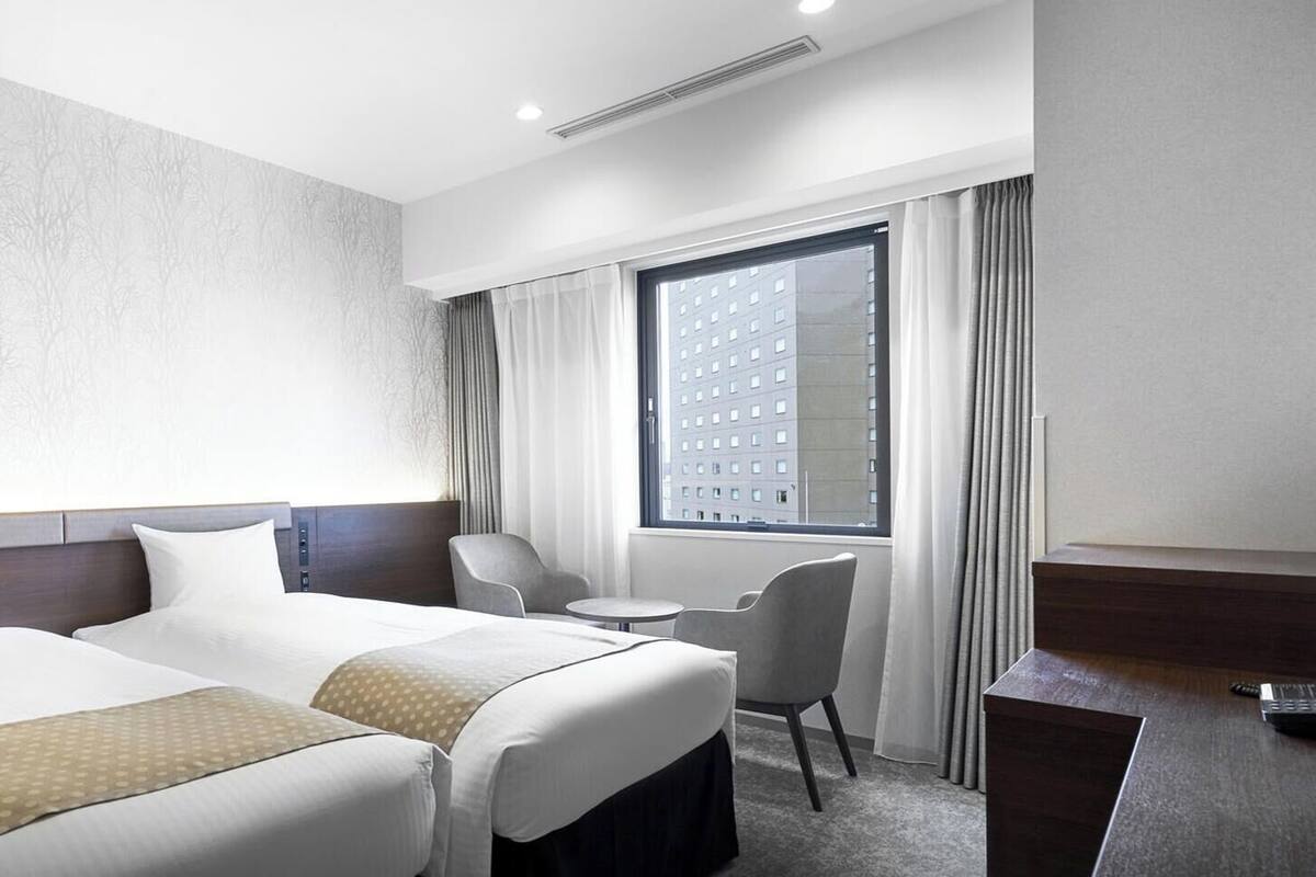 A hotel room with two beds, a desk, a chair, a window with curtains, and a patterned wall.