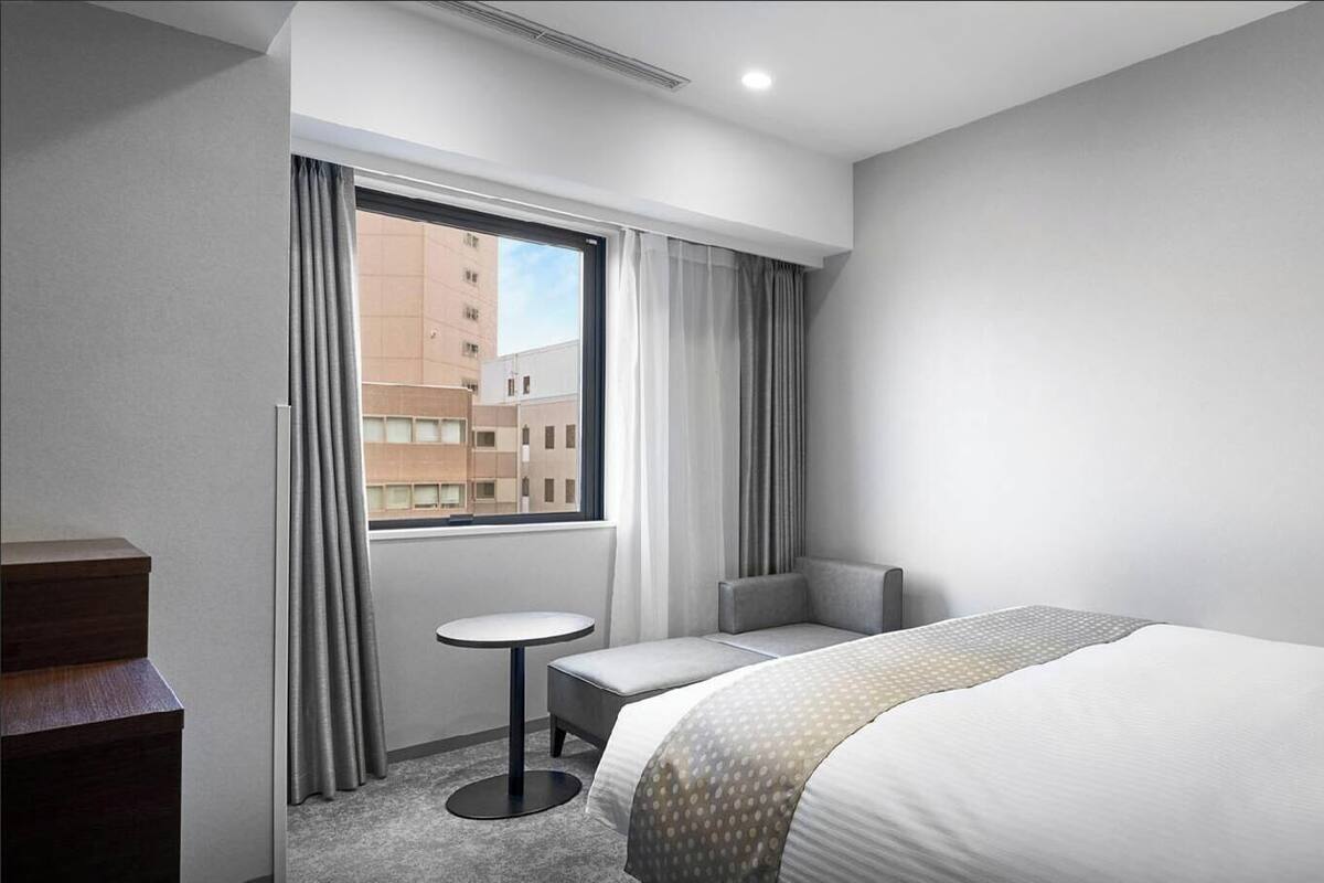 A hotel room with a large bed, a small round table, a chair, a desk, and a window with curtains.