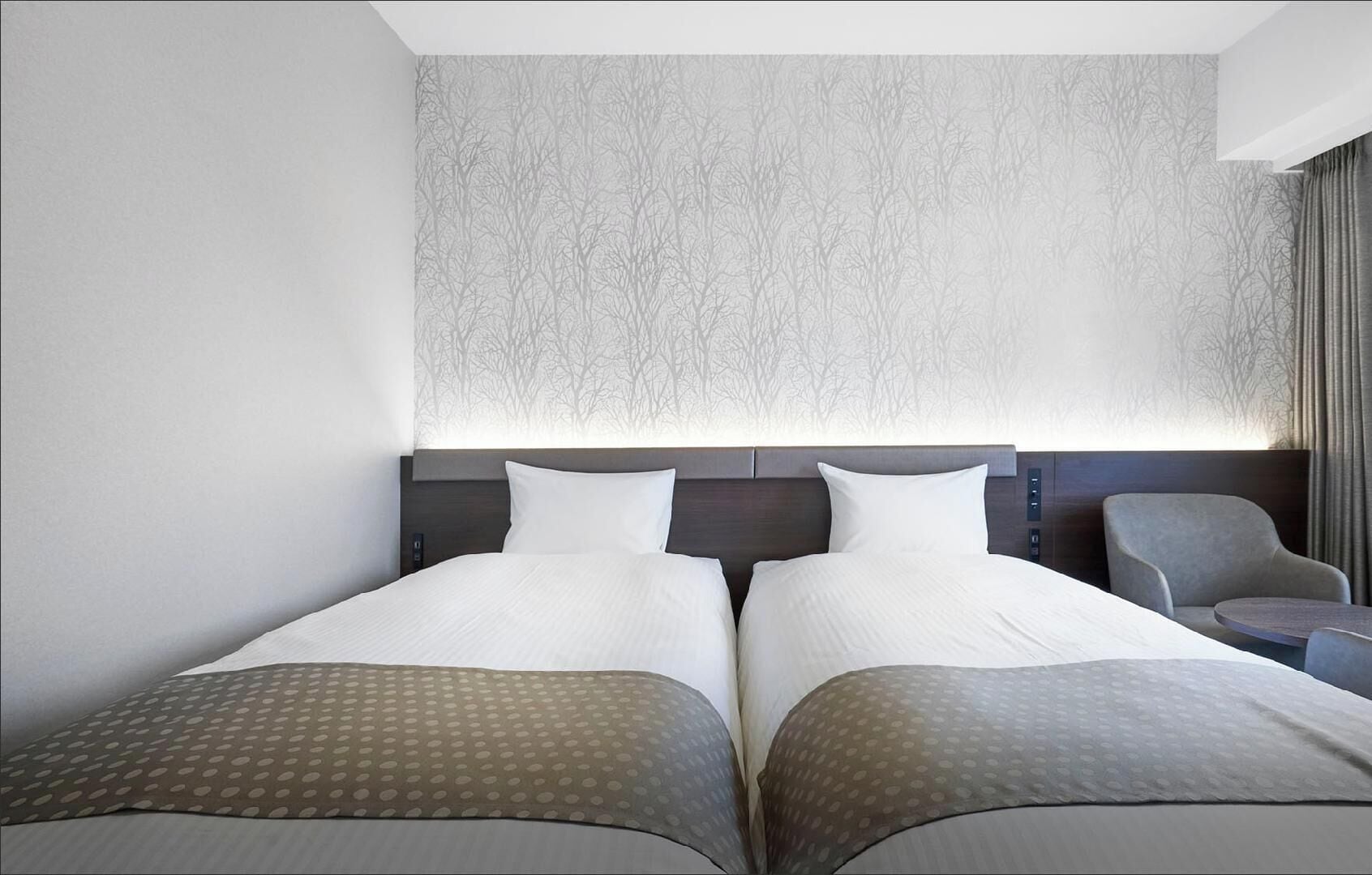A hotel room with two beds, a headboard with control buttons, a chair, and a small table.