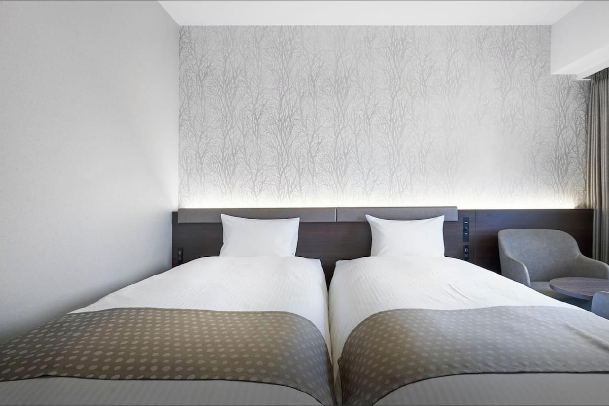 A hotel room with two beds, a headboard with control buttons, a chair, and a small table.