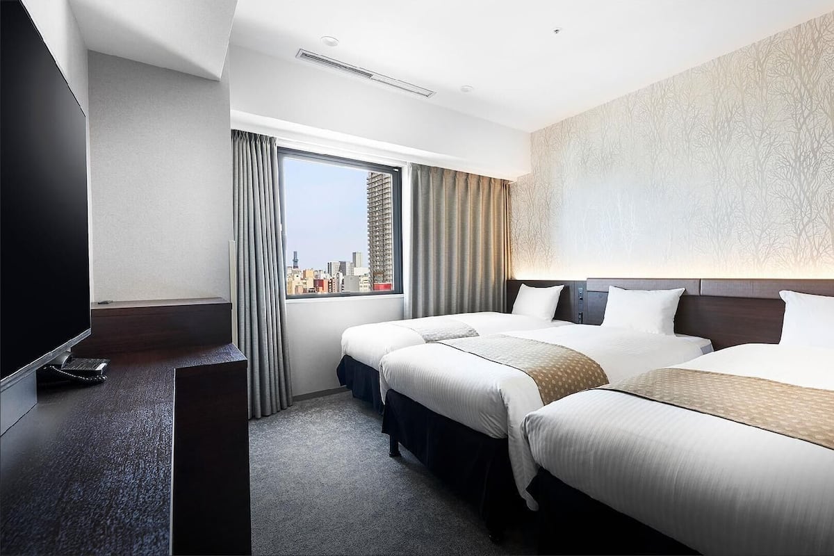 A hotel room with two beds, a TV, a desk, and a large window with a city view.