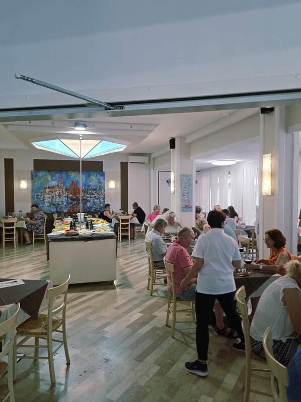 A dining area with multiple tables and chairs, a central buffet, and a large painting on the wall. - Villa Roma (Caorle)